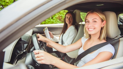Porträt einer lächelnden Person mit offenen, hellen Haaren, die am Steuer eines Autos sitzt. Auf dem Sitz neben ihr sitzt eine weitere Person, die ebenfalls in die Kamera blickt und ein Klemmbrett hält