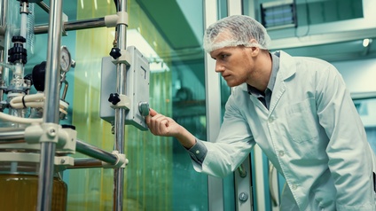 Person in weißem Kittel mit Haarnetz blickt auf Apparatur, an der sie Knopf dreht