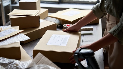 Auf einem Tisch liegen mehrere Pakete, Kuverts sowie durchsichtige Folie. Eine Person steht seitlich vor dem Tisch und hat ihre beiden Händen auf einem Paket