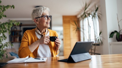 Person mit kurzen hellen Haaren und Brille sitzt freudig mit einer Tasse, einem Notizbuch und einem Tablet bei einem Tisch in einem Wohnraum