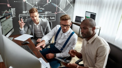 An einem Schreibtisch sitzen drei Personen nebeneinander. Auf dem Tisch sind drei Bildschirme. Zwei der Personen blicken auf einen der Bildschirme. Vor ihnen liegen Zettel. Eine der Personen hält ein Tablet