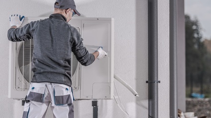 Person in Rückenansicht mit Kappe und Arbeitshandschuhen befestigtWärmepumpen-Außengerät an weißer Hausfassade