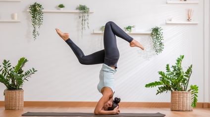Seitliche Aufnahme einer Person in Sportkleidung auf einer Sportmatte in einem hellen Raum mit Topfpflanzen. Die Person macht einen Kopfstand. Sie hat die Hände auf dem Hinterkopf und die Beine angewinkelt