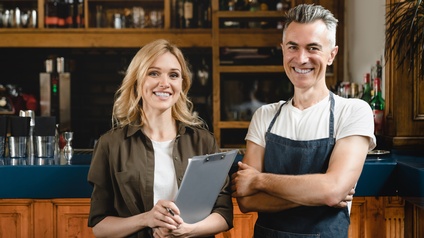 Zwei lächelnde Personen stehen vor der Bar in einem Gastronomiebetrieb, eine Person trägt eine Schürze, die andere hält ein Clipboard in Händen