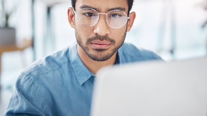 Person mit kurzen dunklen Haaren, Bart und silberner Brille sowie blauem Hemd blickt auf einen Monitor