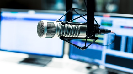 Nahaufnahme eines Mikrofons in einer Nachrichtenstation, im Hintergrund stehen zwei große Monitorbildschirme