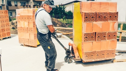 Seitliche Aufnahme einer Person, die mit beiden Händen ein eine niedrige Platte mit Rädern, beladen mit vielen Ziegelsteinen an einer Stange auf einer asphaltierten Unterfläche zieht