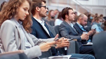 Im Publikumsbereich sitzen viele Personen in mehreren Sesselreihen neben- und hintereinander. Manche Personen blicken auf ihr Handy. Eine hält einen Kaffeebecher. Eine trägt einen Mundschutz.