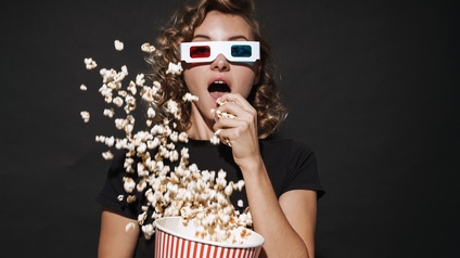 Person mit 3D-Brillen mit einer roten und einer blauen Brillenglasfolie isst Popcorn aus großem Pappbehälter, aus dem Popcorn in die Höhe fliegt