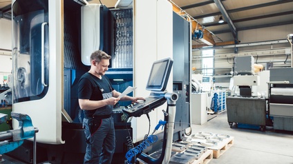 Person steht bei CNC Maschine und liest dabei von einem Zettel in einer Produktionsstätte