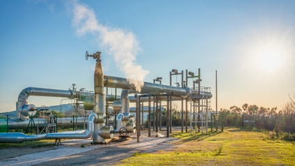 In einem eingezäunten Bereich ist ein Teil asphaltiert. Daneben ist grünes Gras. Auf dem asphaltierten Bereich verlaufen Pipelines in unterschiedlichen Höhen und verschiedenen Breiten. Der Himmel ist blau. An einer Stelle steigt Dampf aus