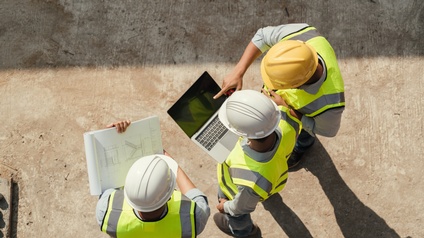 Topshot dreier Personen mit Helmen und Sicherheitswesten, die über eine Baustelle gehen. Die Person links hält einen Plan, die Person in der Mitte einen Laptop und die Person rechts deutet nach vorne mit dem Zeigefinger