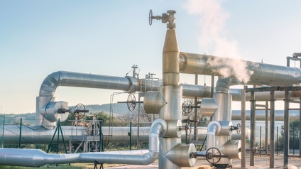 In einem eingezäunten Bereich ist ein Teil asphaltiert. Daneben ist grünes Gras. Auf dem asphaltierten Bereich verlaufen Pipelines in unterschiedlichen Höhen und verschiedenen Breiten. Der Himmel ist blau. An einer Stelle steigt Dampf aus