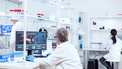 Personen in weißen Kitteln sitzen mit Schutzhandschuhen und einer Schutzbrille im Labor und untersuchen Virenbilder auf dem Computer. Im Labor befinden sich unzählige Probegefäße und Laborutensilien.