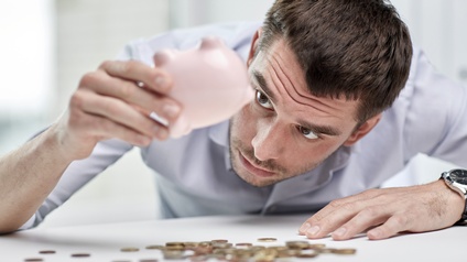 Person mit kurzen dunklen Haaren blickt erwartungsvoll auf ein ausgeleertes  Sparschwein während Münzen auf dem Tisch liegen