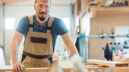 Porträt einer Person mit Schutzbrille, Latzhose, kurzen, hellen Haaren und Bart, die ein Schleifpapier auf ein Holzbrett hält