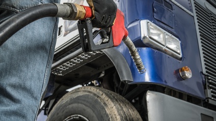 Detailansicht eines rotschwarzen Zapfhahnes, der von Hand mit Handschuh gehalten wird, im Hintergrund im Ausschnitt blauer Lastkraftwagen