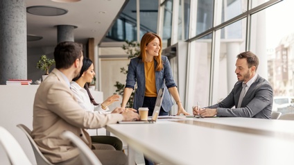 An einem langen Tisch in einem großen Raum mit einer Glasfront sitzen drei Personen. Eine Person steht am Tisch. Eine der sitzenden Personen hat einen aufgeklappten Laptop vor sich. Eine andere Person notiert etwas auf einem Notizblock