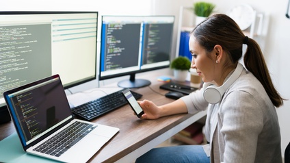 Seitliche Aufnahme einer Person, die an einem Schreibtisch sitzt. Auf dem Tisch sind zwei Bildschirme, eine Tastatur und ein aufgeklappter Laptop. Auf allen sind Quellcodes. Die Person hält ein Smartphone, auf das sie blickt