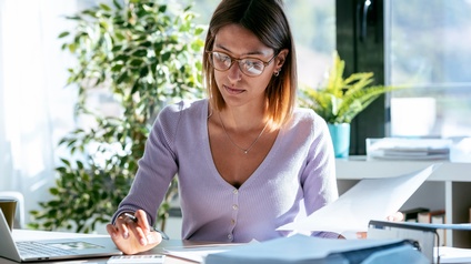Eine Person mit Brille und kinnlangen Haaren sitzt an einem Tisch. In der linken Hand hält sie einen Zettel. Mit der rechten Hand tippt sie auf einem Taschenrechner. Auf dem Tisch sind weitere Unterlagen und ein Laptop