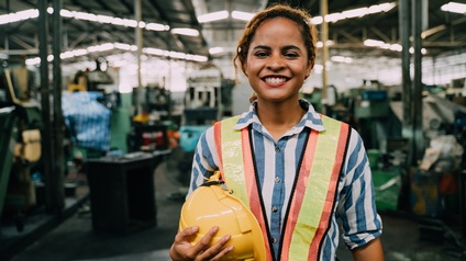 Halbporträt einer Person mit Sicherheitsweste und Helm in der rechten Hand. Im Hintergrund ist eine hohe Halle mit Geräten