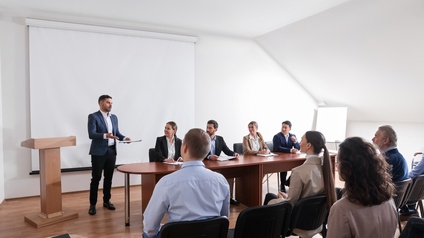 Personen in Businesskleidung sitzen in einem Besprechungsraum zusammen, eine Person steht und hält eine Rede