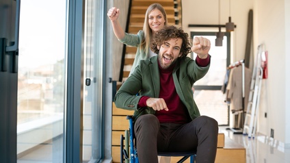 Zwei junge Personen befinden sich in einem modernen hellen Raum, lächeln und strecken eine Faust zur Kamera, die vordere Person sitzt in einem Rollstuhl