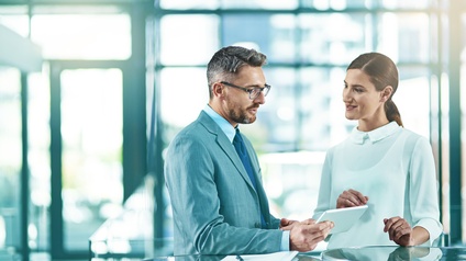 Zwei Personen stehen an Tischen in Unterhaltung, eine Person hält Tablet in Händen, am Tisch Dokument mit Stift liegend
