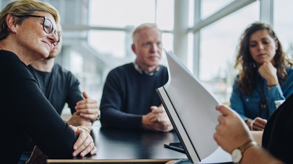 Mehrere Personen sitzen an Konferenztisch, Ausschnitt einer Person, die in Dokumentenmappe blättert 