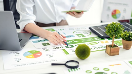 Eine Person sitzt an einem Schreibtisch. Mit der rechten Hand hält sie einen Stift und deutet auf einen Zettel, der vor ihr liegt. Daneben sind weitere Zettel. Auf den Zettel sind verschiedene Diagramme und Icons. Darauf liegt ein kleiner, grüner Ball