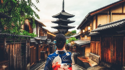 Rückenansicht einer Person in einem blau-weiß-rotem Oberteil mit großen Blumen sowie einer hochgesteckten Frisur, die auf einer Straße durch einen Ort geht. Links und rechts sind einstöckige Gebäude