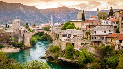 Über einen Fluss führt eine hohe Brücke aus Stein. Links und rechts davon sind leicht abfallend mehrere verschiedene Gebäude. Manche sind aus Stein. Im Hintergrund ist eine Bergkette