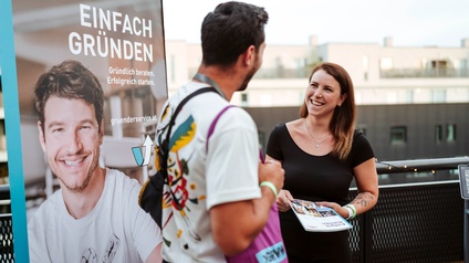 2 Personen stehen auf einer Terrasse vor einem Rollup mit der Aufschrift 