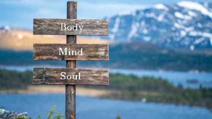 Holzwegtafel mit der Beschriftung Body, Mind, Soul, im Hintergrund verschwommen Seenlandschaft mit beschneiter Bergkette