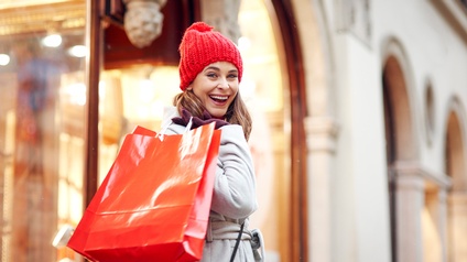 Person mit roter Strickmütze und grauem Mantel hält rote Einkaufstaschen über der rechten Schulter und blickt freudig in die Kamera, im Hintergrund zeigt sich eine Fassade einer Straße