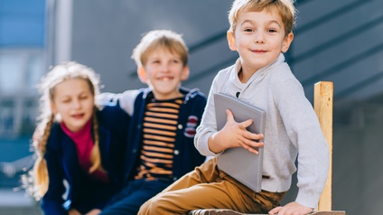 Fröhliches Kind mit Buch sitzt auf einer Bank und blickt in die Kamera während im Hintergrund zwei weitere glückliche Kinder sitzen