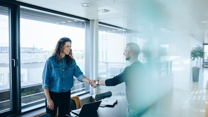 Zwei lächelnde Personen reichen sich in Besprechungsraum über Tisch hinweg die Hand
