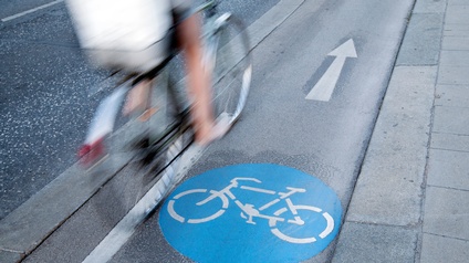 Asphaltstraße mit weißem aufgemalten Fahrradzeichen und einem weißen Pfeil, auf dem eine Person auf einem Fahrrad fährt. Links neben dieser Straße ist eine weitere mehrspurige Straße. Rechts daneben ist ein Gehweg