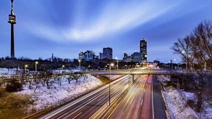 Aufnahme einer mehrspurigen Straße aus der Vogelperspektive. Links und rechts von der Straße liegt Schnee. Über die Straße verläuft eine Autobrücke. Im Hintergrund sind mehrere Wolkenkratzer. Links im Bild ist ein Turm