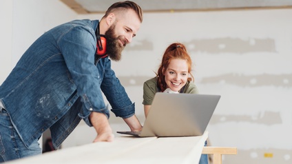 Zwei lächelnde Personen blicken auf vor sich aufgeklappten Laptop, eine Person trägt roten Gehörschutz um den Hals, im Hintergrund verschwommen rohverputzte Wand