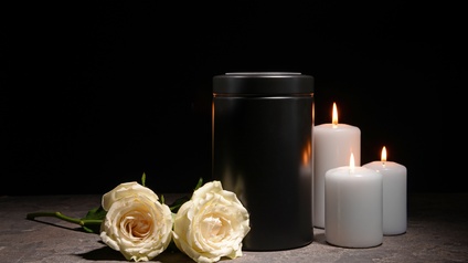 Schwarze Urne, nebenliegend zwei weiße Rosen und drei brennende weiße Kerzen, Hintergrund schwarz