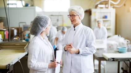 Zwei Personen in weißer Schutzbekleidung mit Haarnetzen unterhalten sich in Lebensmittelproduktionsstätte stehend