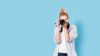 Frontale Aufnahme einer Person vor einer blauen Wand, die einen blonden Dutt hat und mit einer Kamera fotografiert. Die Person trägt eine Jeans, ein weißes Oberteil und ein hellblaues, geöffnetes Hemd