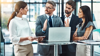 Vier Personen in Bürokleidung stehen nebeneinander an einem Stehtisch, auf dem ein Laptop steht. Die Person am Laptop gestikuliert mit den Händen, die anderen blicken zu ihr. Im Hintergrund sind Fensterfronten