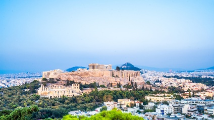 Panoramaaufnahme von Athen mit der Akropolis im Zentrum. Rundherum sind grüne Bäume so wie sehr viele Wohngebäude. Im Hintergrund sind Hügel
