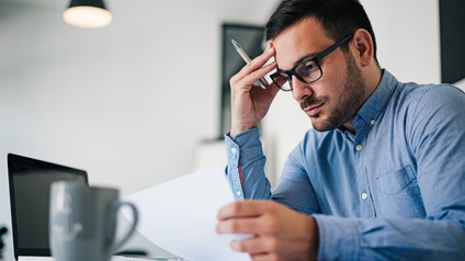 Seitliche Aufnahme einer Person mit Hemd, Brille, kurzen, dunklen Haaren und Bart, die an einem Tisch sitzt und auf einen Zettel blickt, den sie hält. Auf dem Tisch sind weitere Zettel, ein Laptop und eine Tasse