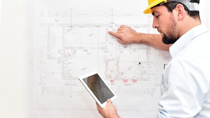 Person mit gelbem Helm deutet mit Finger auf Gebäudeplan auf Wand und hält in anderer Hand Tablet