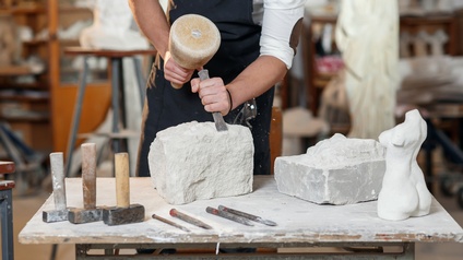 Eine Person mit Schürze im Ausschnitt, die mit Hammer und Meißel einen Stein bearbeitet. Auf dem Arbeitstisch befinden sich Steine, eine Steinskulptur, Hammer und Meißel. Dahinter erkennt man eine Werkstatt.