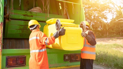 Zwei Personen in gelben Schutzhelmen mit orangen Warnwesten in Rückenansicht beim Ausleeren einer gelben Mülltonne in grünen Müllabfuhrwagen