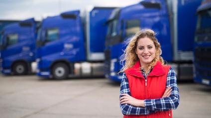 Portrait einer lächelnde Person in roter Weste, die Arme verschränkt vor Körper hält, im Hintergrund verschwommen blaue parkende Lastkraftwagen